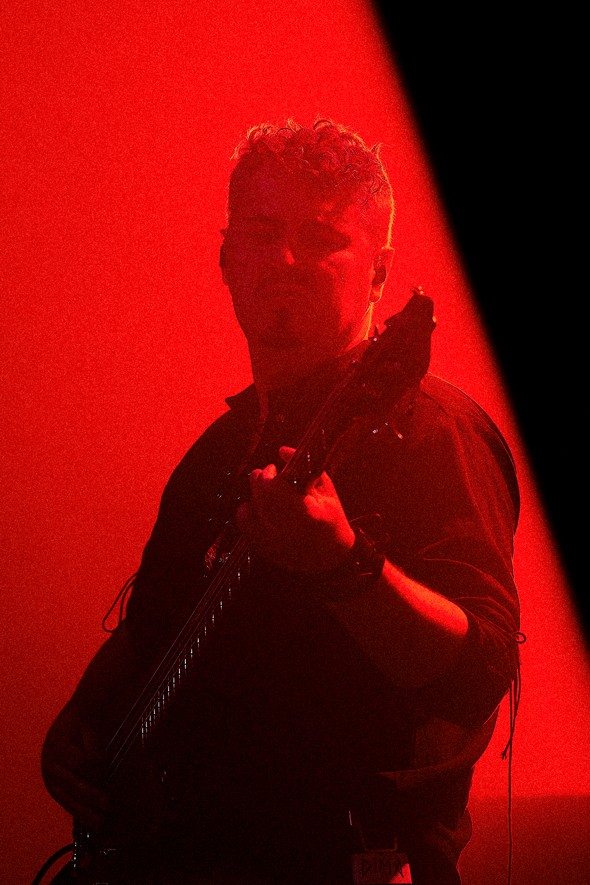 Basista na scenie oświetlony intensywnym czerwonym światłem, gra na gitarze basowej, częściowo pogrążony w cieniu, co tworzy mroczny, koncertowy klimat.