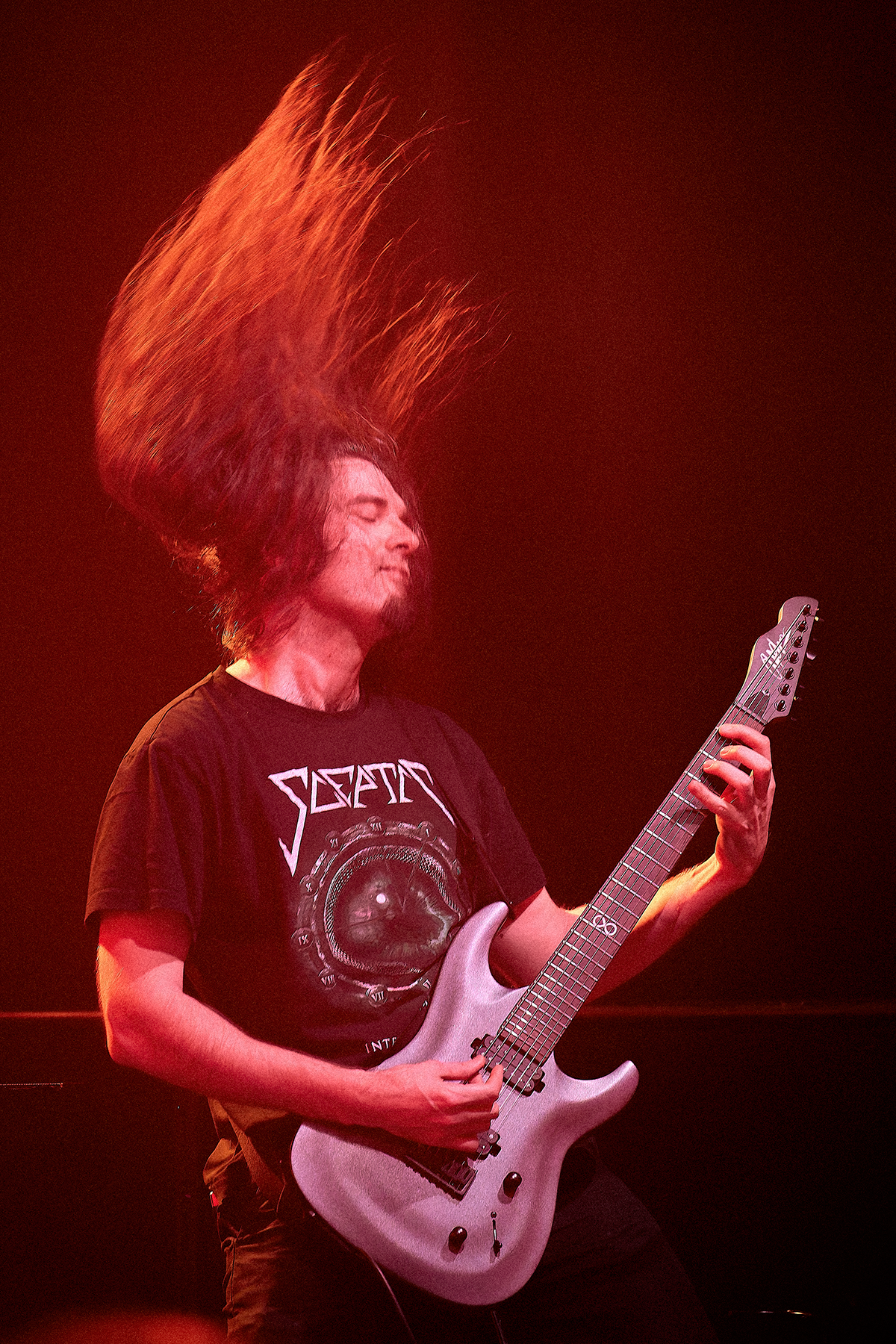 Gitarzysta podczas koncertu, oświetlony czerwonym światłem, gra na gitarze elektrycznej, wykonując energiczny headbanging z włosami uniesionymi w powietrzu.