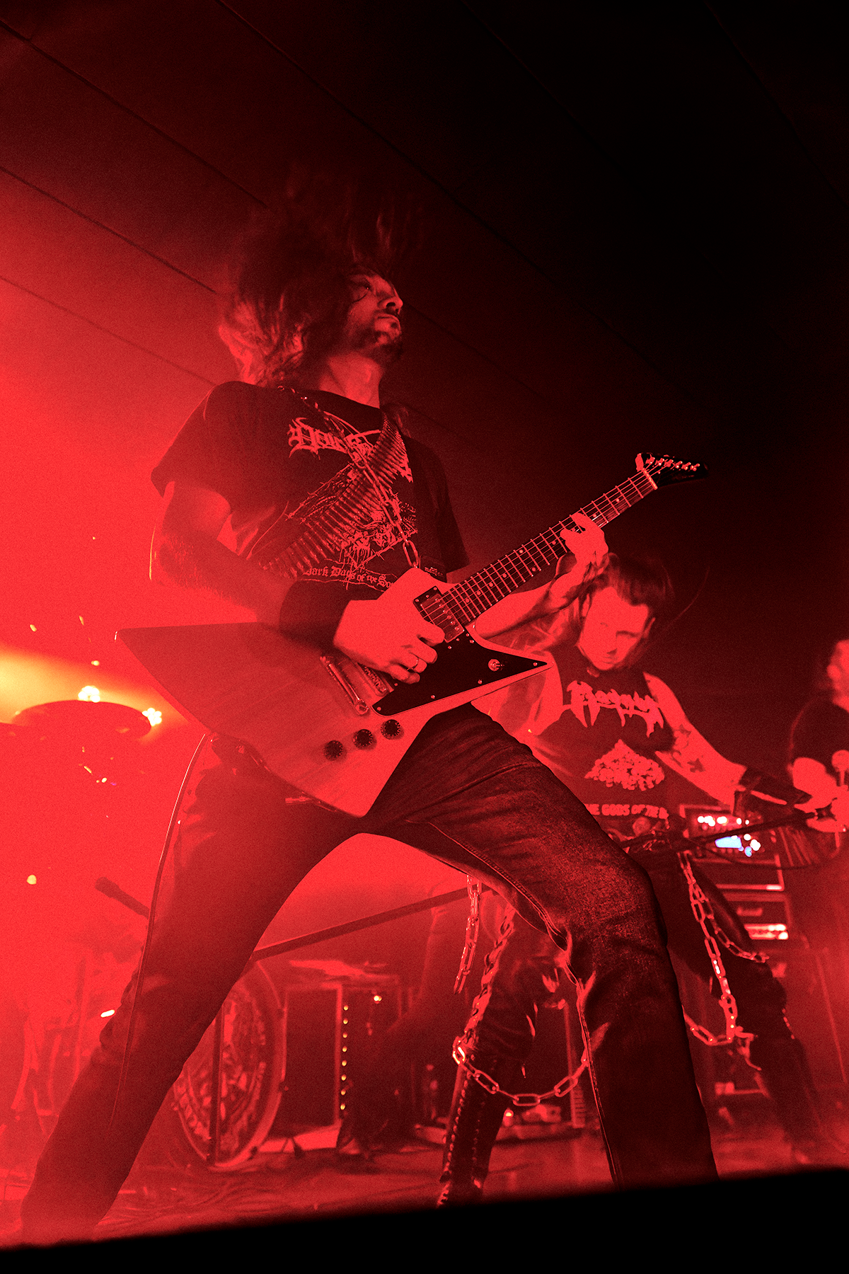 Gitarzysta zespołu Tribute to Magnus gra na scenie w intensywnym czerwonym świetle, z uniesioną nogą i rozwianymi włosami; w tle drugi muzyk i perkusja, tworząc surowy, deathmetalowy klimat koncertu.