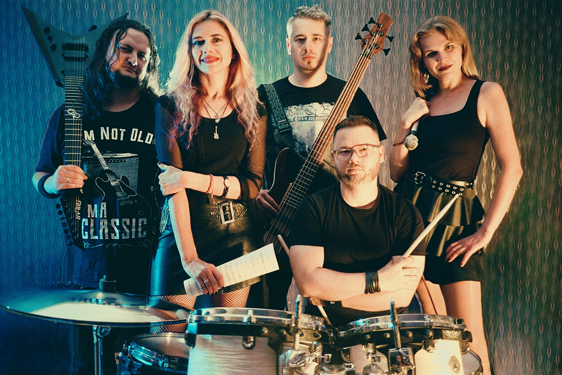 Pięcioosobowy zespół muzyczny pozuje razem w pomieszczeniu prób — dwie wokalistki z mikrofonem i nutami, gitarzysta z gitarą elektryczną, basista oraz perkusista siedzący przy zestawie perkusyjnym. Całość utrzymana w klimatycznym, lekko retro oświetleniu.