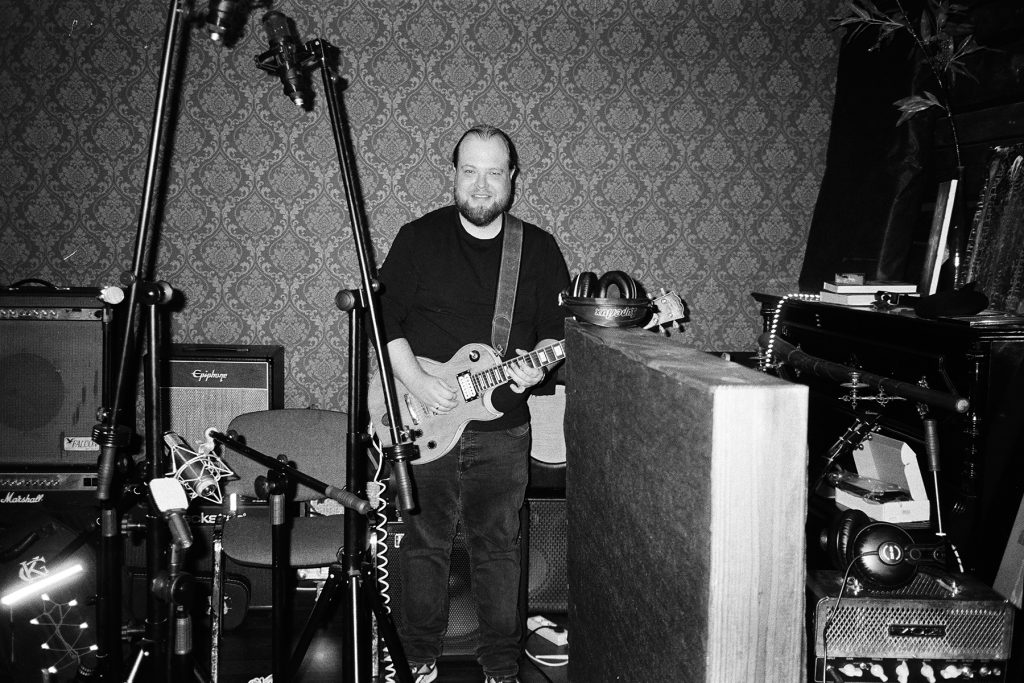 Czarno-białe zdjęcie przedstawiające Ambro z Mama Morphine podczas sesji nagraniowej w Jupiter Ranch Studio. Muzyk stoi z gitarą elektryczną pośród mikrofonów, wzmacniaczy i sprzętu studyjnego, uśmiechając się w stronę aparatu. Pomieszczenie ma surowy, analogowy klimat, a fotografia oddaje atmosferę pracy nad nagraniami oraz rock’n’rollowy charakter sesji.