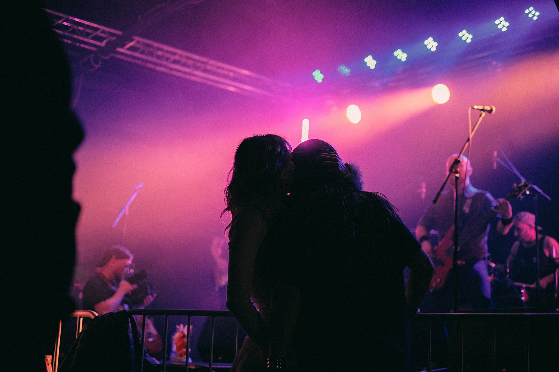 Sylwetka całującej się pary stojącej pod sceną podczas Wajbzzfest! 2025, gdy w tle koncert gra Mama Morphine. Scena skąpana jest w fioletowym i różowym świetle, a rozmyte sylwetki muzyków podkreślają koncertowy klimat zdjęcia. Fotografia oddaje emocjonalną i intymną atmosferę wydarzenia oraz energię muzyki na żywo.