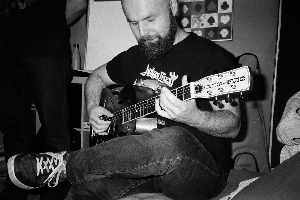 Czarno-białe zdjęcie przedstawiające Dynię z Mama Morphine siedzącego podczas sesji nagraniowej i grającego na gitarze. Muzyk ma założoną koszulkę zespołu Judas Priest i skupia się na instrumencie trzymanym na kolanach. Fotografia została wykonana w swobodnej atmosferze studia nagraniowego i oddaje luźny, naturalny klimat pracy nad muzyką.