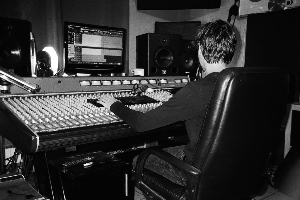 Czarno-białe zdjęcie przedstawiające Pitera z Jupiter Ranch Studio siedzącego przy dużej analogowej konsoli mikserskiej podczas sesji nagraniowej Mama Morphine. Realizator skupia się na pracy przy mikserze i komputerze wyświetlającym ślady nagraniowe. W studiu widoczne są monitory odsłuchowe, mikrofon oraz sprzęt audio, a całość utrzymana jest w surowym, dokumentalnym klimacie oddającym atmosferę pracy nad muzyką.