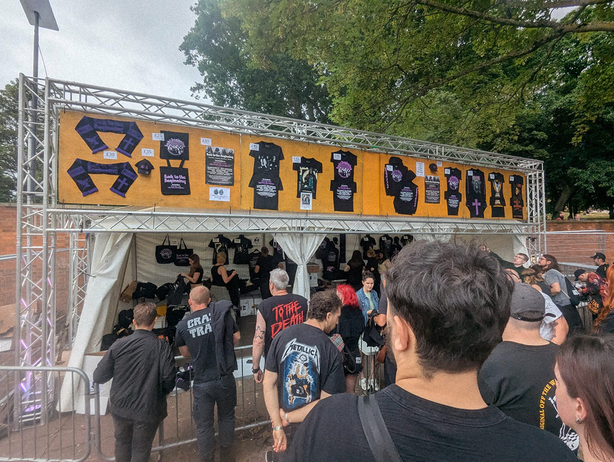 Stoisko z merchandisem koncertowym na świeżym powietrzu, z wyeksponowanymi koszulkami i gadżetami „Back to the Beginning”, przed którym ustawia się kolejka fanów ubranych w czarne, metalowe t-shirty.
