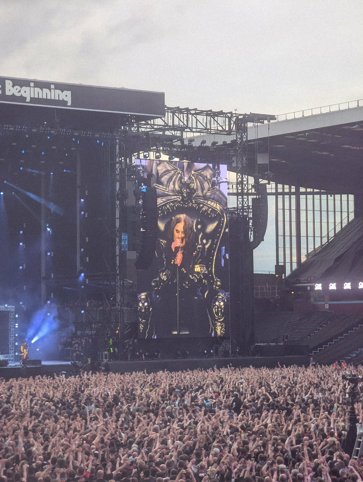 Ogromny tłum fanów na stadionie podczas koncertu, z uniesionymi rękami skierowanymi w stronę sceny, gdzie na wielkim ekranie widać Ozzy’ego Osbourne’a siedzącego na charakterystycznym tronie i śpiewającego do mikrofonu.