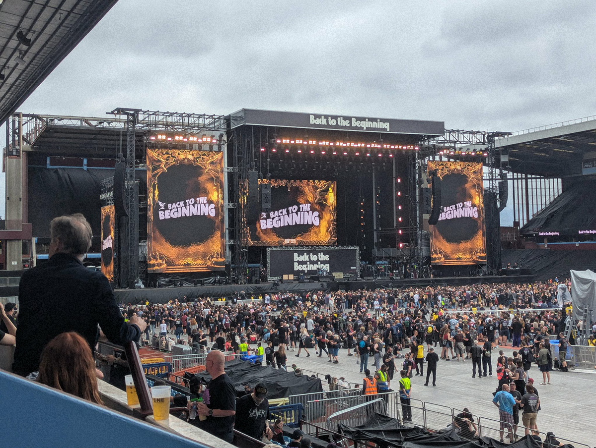 Widok na ogromną scenę koncertową „Back to the Beginning” na stadionie, z wielkimi ekranami i tłumem fanów gromadzących się przed rozpoczęciem występów.