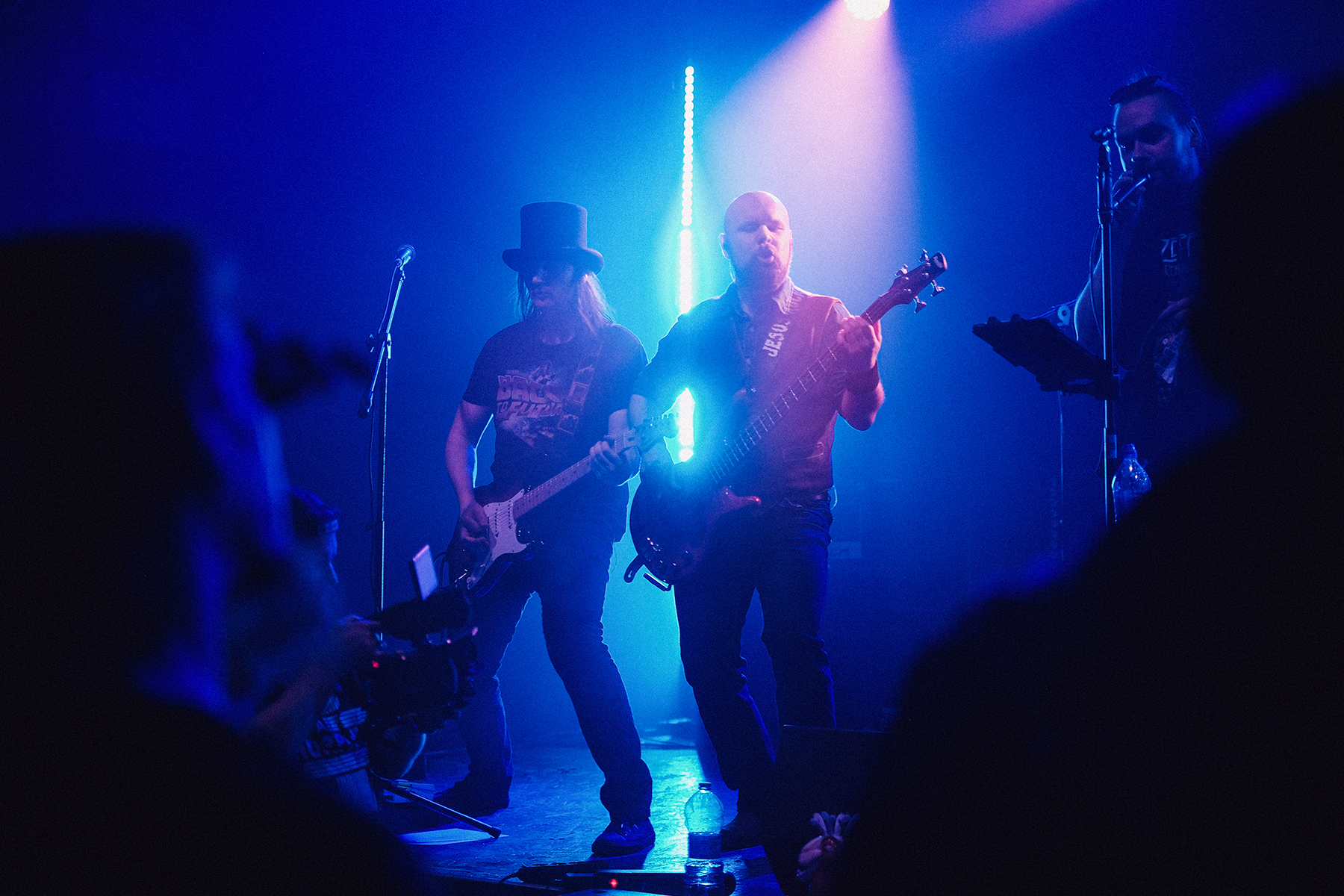 Muzycy wrocławskiego zespołu Mama Morphine podczas koncertu na Wajbzzfest! 2025 grają na scenie oświetlonej intensywnym niebiesko-fioletowym światłem. Gitarzysta w charakterystycznym cylindrze i basista stoją obok siebie w centrum sceny, a po lewej stronie widoczny jest operator kamery z Wajbzz rejestrujący występ. Zdjęcie oddaje energię i klimat rockowego koncertu na żywo.