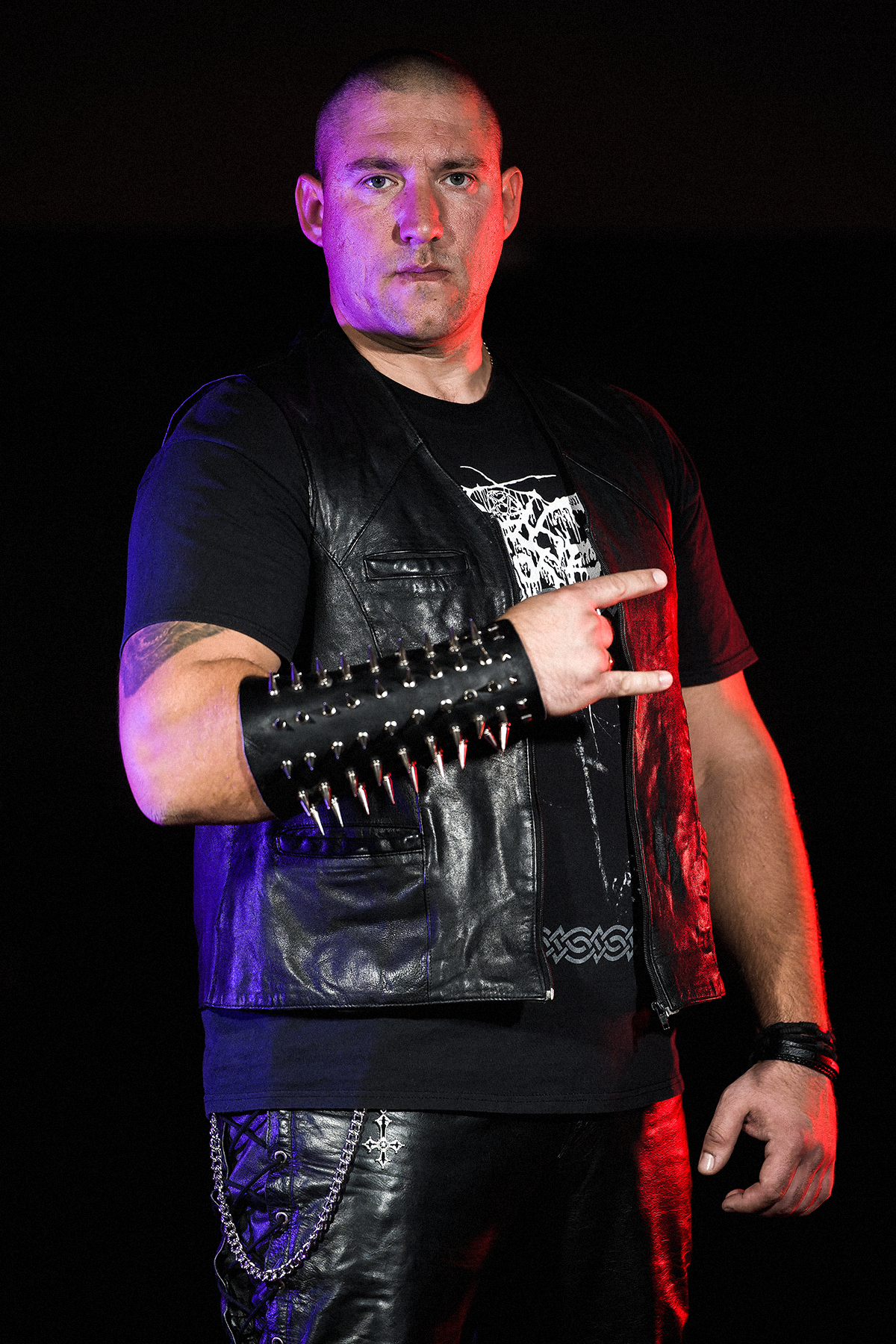 Portret muzyka black metalowego pozującego na ciemnym tle z gestem „rogów”. Mężczyzna ubrany jest w czarną skórzaną kamizelkę, metalowe naramienniki z ćwiekami oraz czarne sceniczne ubrania. Zdjęcie oświetlone jest czerwonym i fioletowym światłem, podkreślającym mroczny klimat fotografii.