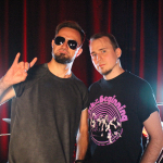 Dwóch muzyków wrocławskiego zespołu Slavemind — gitarzysta i perkusista — stoi na sali prób. Gitarzysta w okularach przeciwsłonecznych pokazuje znak „rock on”, a perkusista ma na sobie koszulkę z motywem koncertu „Back to the Beginning”. W tle widać zestaw perkusyjny i czerwone oświetlenie. Zdjęcie promuje odcinek podcastu Wajbzz o jego wyjeździe na to wydarzenie.