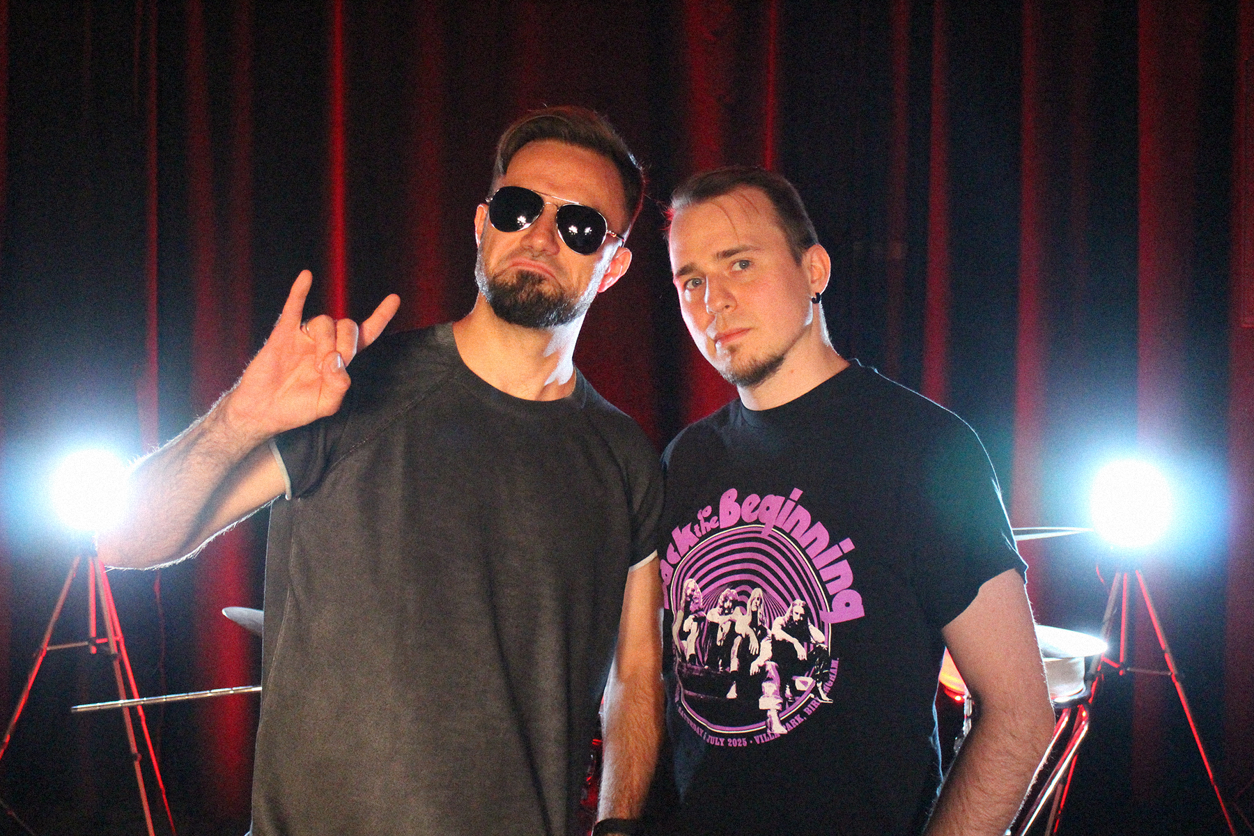 Dwóch muzyków wrocławskiego zespołu Slavemind — gitarzysta i perkusista — stoi na sali prób. Gitarzysta w okularach przeciwsłonecznych pokazuje znak „rock on”, a perkusista ma na sobie koszulkę z motywem koncertu „Back to the Beginning”. W tle widać zestaw perkusyjny i czerwone oświetlenie. Zdjęcie promuje odcinek podcastu Wajbzz o jego wyjeździe na to wydarzenie.