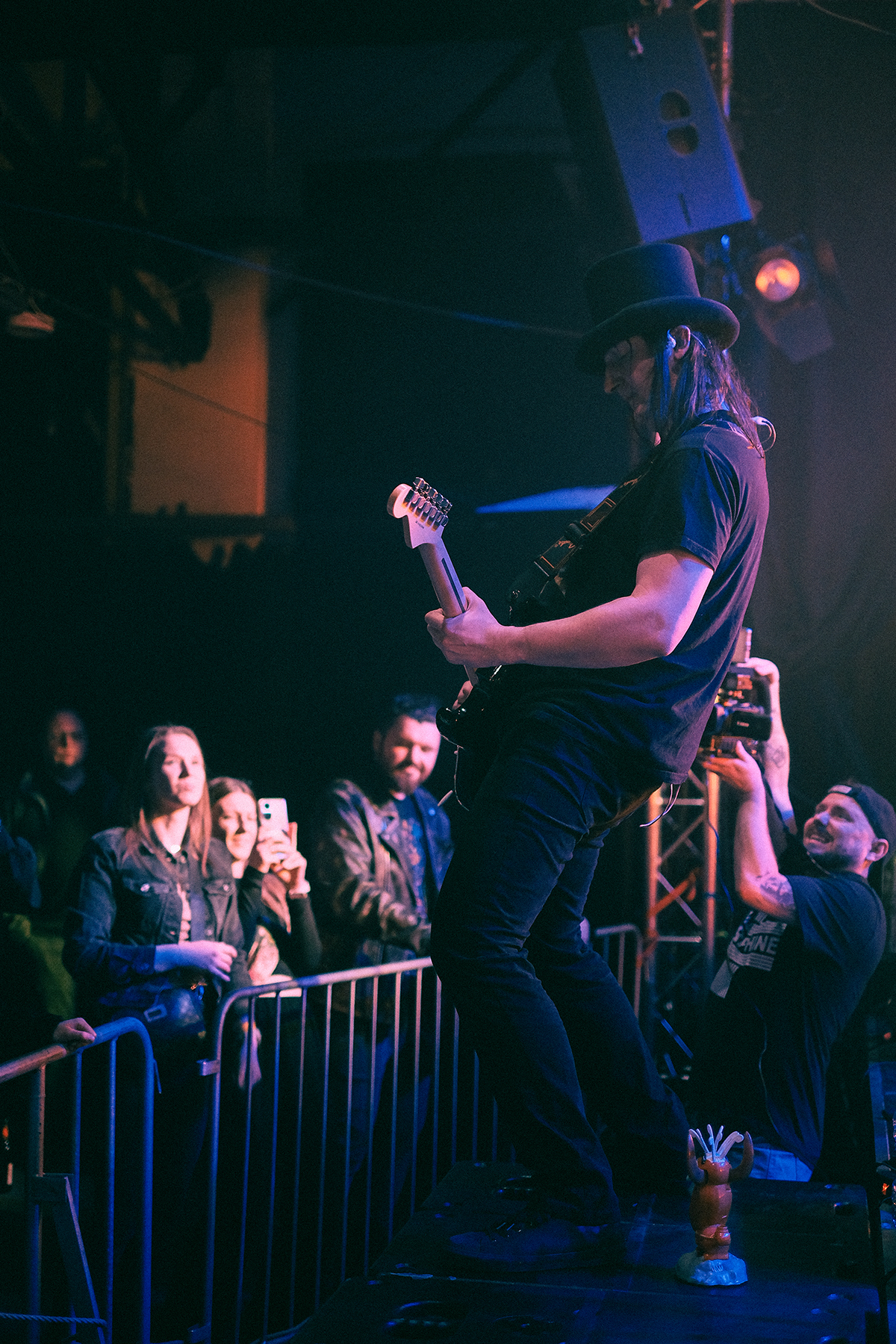 Gitarzysta wrocławskiego zespołu Mama Morphine podczas koncertu na Wajbzzfest! 2025 stoi na scenicznej kolumnie z gitarą elektryczną. Muzyk ubrany jest na czarno i ma charakterystyczny cylinder, a pod sceną widoczna jest publiczność oraz operator kamery z Wajbzz rejestrujący występ. Zdjęcie utrzymane jest w koncertowym klimacie z ciemnym, niebiesko-fioletowym oświetleniem.