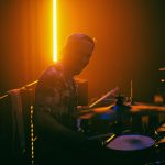 Perkusista wrocławskiego zespołu Mama Morphine podczas występu na Wajbzzfest! 2025. Muzyk siedzi za zestawem perkusyjnym, oświetlony intensywnym pomarańczowym światłem scenicznym, a w tle widoczny jest pionowy pasek LED. Zdjęcie utrzymane jest w mrocznym, koncertowym klimacie z mocnym kontrastem i ciepłą kolorystyką.