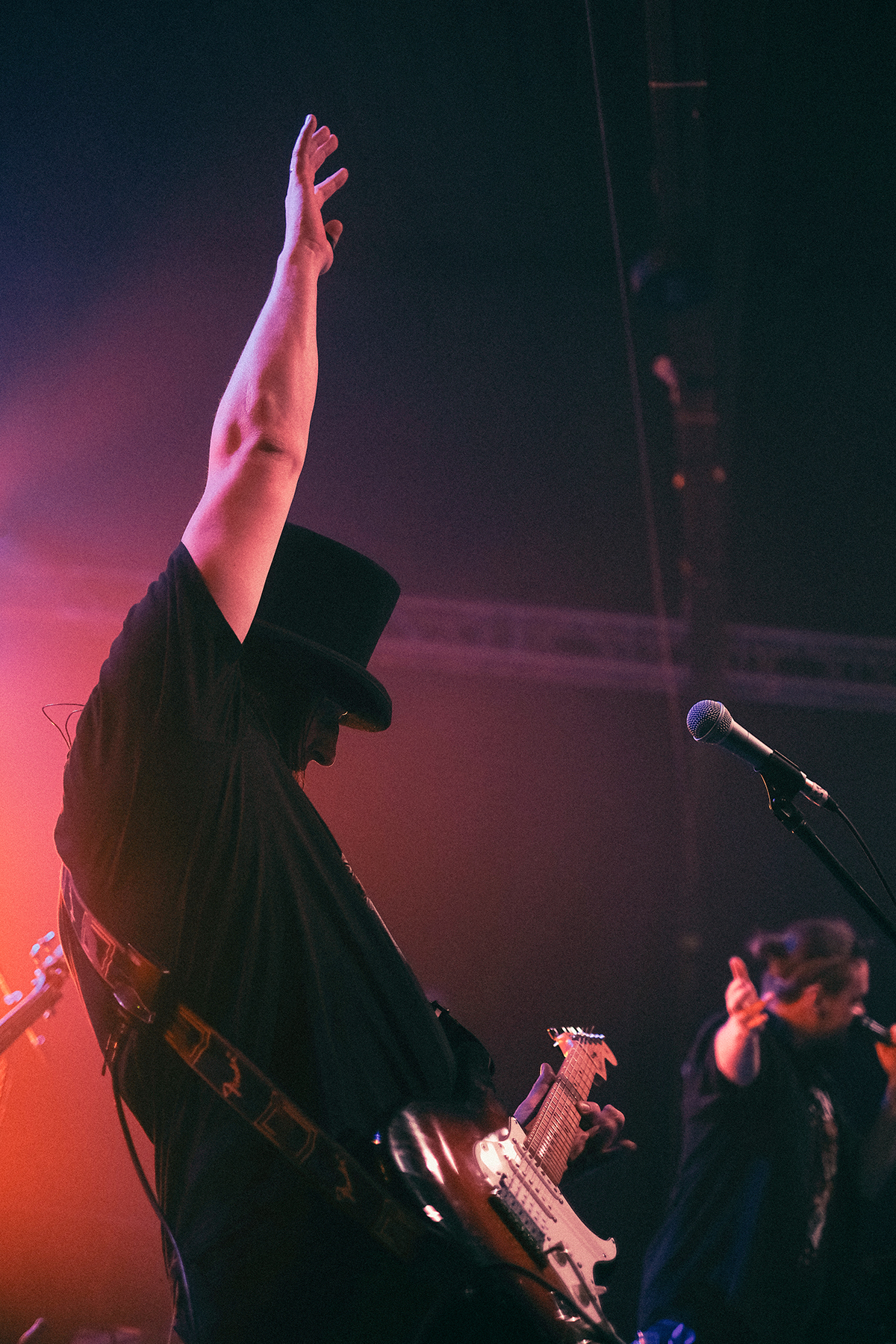 Gitarzysta wrocławskiego zespołu Mama Morphine podczas koncertu na Wajbzzfest! 2025 unosi rękę w górę, trzymając gitarę elektryczną na scenie skąpanej w czerwono-różowym świetle. Muzyk ma charakterystyczny cylinder i ubrany jest na czarno, a w tle widoczny jest wokalista zespołu. Zdjęcie oddaje dynamiczny, rockowy klimat koncertu na żywo.