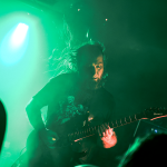 Gitarzysta zespołu Tribute to Magnus gra na scenie w zielonym świetle, wykonując utwory inspirowane polskim death metalem lat 80.; dynamiczna poza, rozwiane włosy i publiczność w cieniu na pierwszym planie.