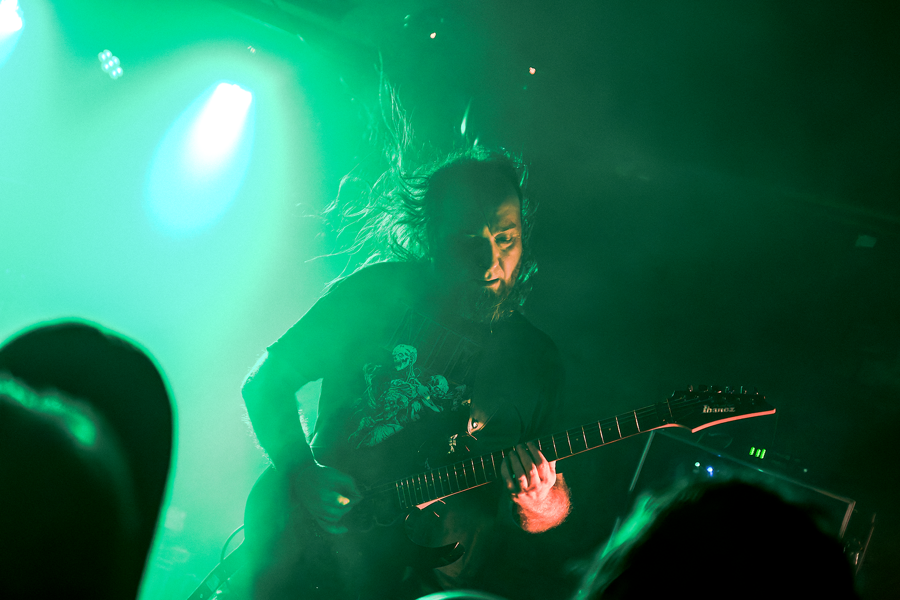 Gitarzysta zespołu Tribute to Magnus gra na scenie w zielonym świetle, wykonując utwory inspirowane polskim death metalem lat 80.; dynamiczna poza, rozwiane włosy i publiczność w cieniu na pierwszym planie.