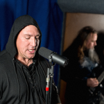 Wokalista w czarnej bluzie z kapturem śpiewający do mikrofonu w sali prób. W tle rozmyty gitarzysta grający na gitarze elektrycznej, całość utrzymana w mrocznym klimacie metalowym.