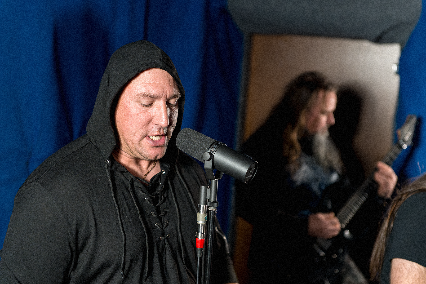 Wokalista w czarnej bluzie z kapturem śpiewający do mikrofonu w sali prób. W tle rozmyty gitarzysta grający na gitarze elektrycznej, całość utrzymana w mrocznym klimacie metalowym.