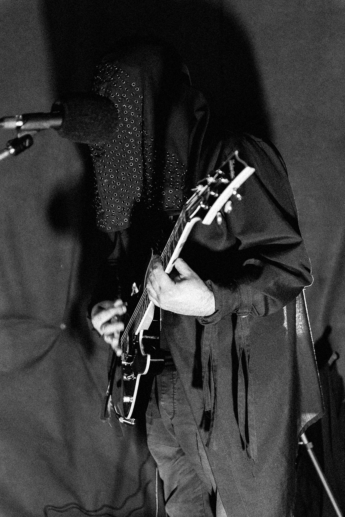 Czarno-biała fotografia gitarzysty podczas koncertu black metalowego. Muzyk ubrany jest w mroczny sceniczny strój z peleryną i zasłoniętą twarzą pokrytą metalowymi nitami. Trzyma gitarę elektryczną i stoi przy mikrofonie w ciemno oświetlonej przestrzeni koncertowej.