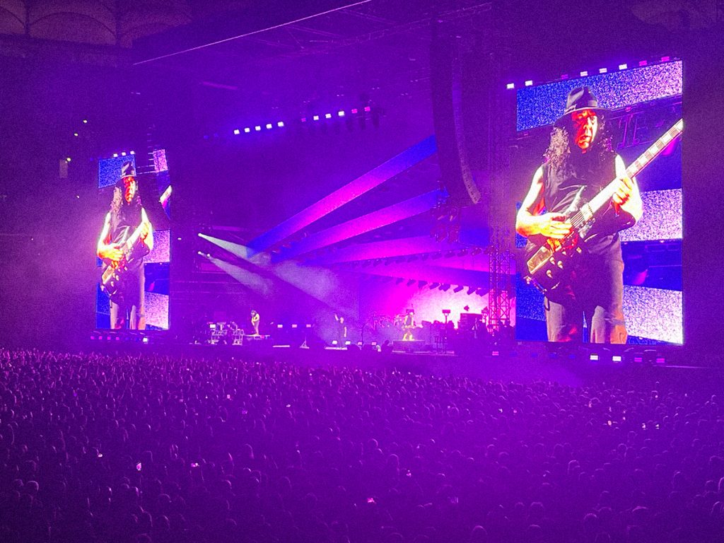 Daron Malakian, gitarzysta i wokalista System of a Down, gra na scenie PGE Narodowego w Warszawie 18 lipca 2026 roku. Muzyk jest wyświetlany na ogromnych telebimach podczas koncertu przed dziesiątkami tysięcy fanów.
