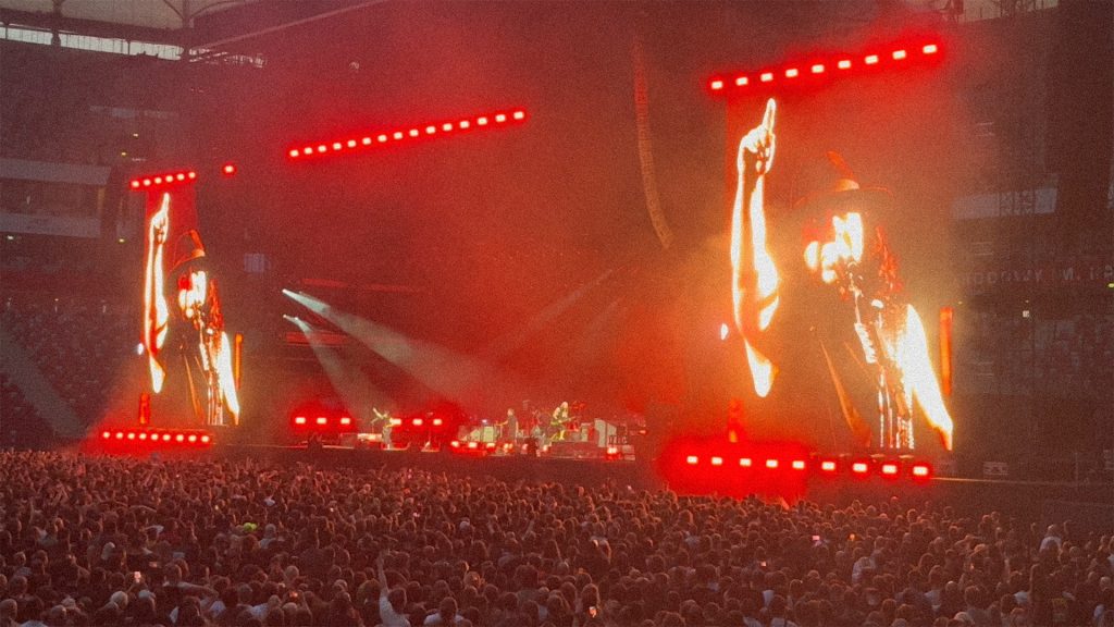 Daron Malakian, gitarzysta i wokalista zespołu System of a Down, występuje na scenie PGE Narodowego w Warszawie 18 lipca 2026 roku. Muzyk jest widoczny na ogromnych telebimach podczas koncertu przed tysiącami fanów.