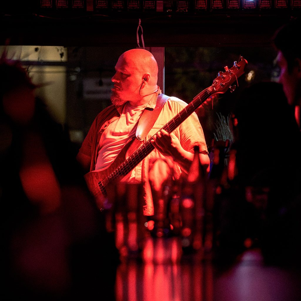 Gibber, basista wrocławskiego zespołu GMOH, gra koncert w klubie Alive pod nasypem we Wrocławiu. Czerwone światła sceniczne podkreślają mroczny klimat występu, a skupiona postawa muzyka i publiczność na pierwszym planie oddają energię koncertu blackened death metalowego zespołu.