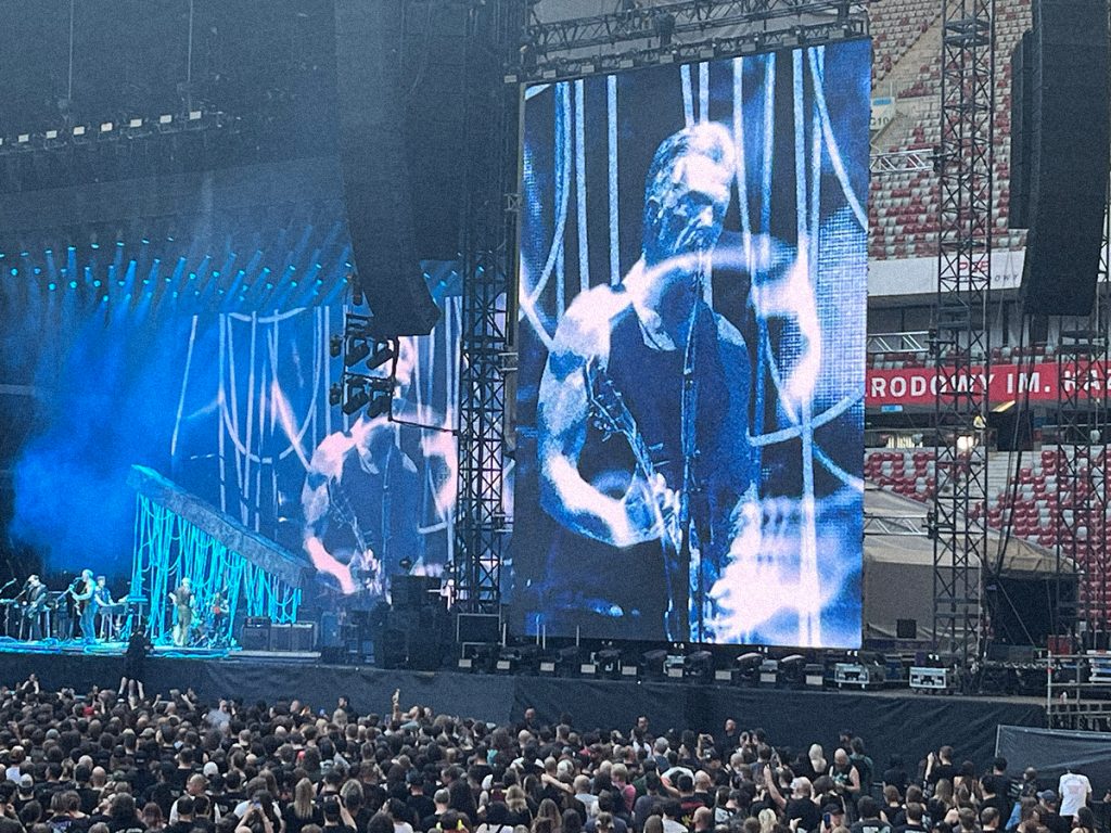 Josh Homme, wokalista i gitarzysta zespołu Queens of the Stone Age, występuje na scenie PGE Narodowego w Warszawie 18 lipca 2026 roku. Muzyk jest widoczny na ogromnym ekranie podczas koncertu poprzedzającego występ System of a Down.