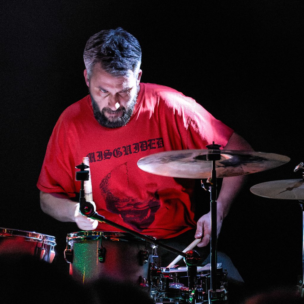 Krzysztof, perkusista zespołu GMOH, gra podczas koncertu w klubie Alive we Wrocławiu. Muzyk w czerwonej koszulce jest oświetlony punktowym światłem i z pełnym skupieniem uderza w werbel oraz tomy zestawu perkusyjnego. Ciemne tło i kontrastowe światło podkreślają intensywny, koncertowy klimat występu.