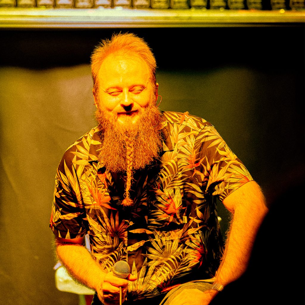 Marcin, wokalista GMOH, podczas koncertu w klubie Alive we Wrocławiu. Muzyk z uśmiechem trzyma mikrofon w dłoni, a ciepłe światło sceniczne podkreśla kameralny charakter chwili między kolejnymi fragmentami występu.