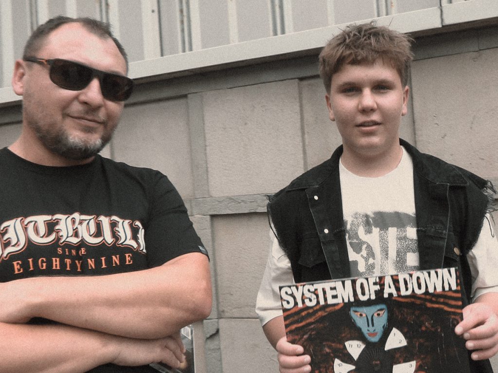Olek, 16-letni fan System of a Down, wraz ze swoim tatą Krzyśkiem pozują do zdjęcia przed koncertem zespołu. Olek trzyma płytę winylową „System of a Down”, a Krzysiek od lat wspiera syna w jego pasji do muzyki metalowej.