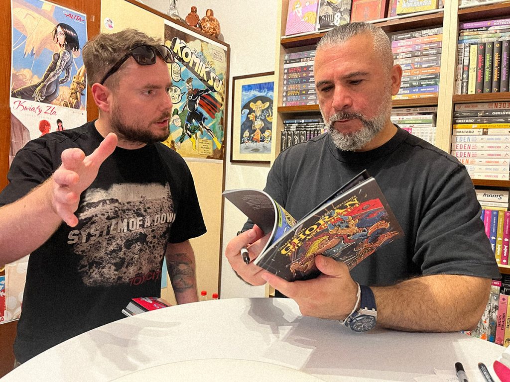 Bartłomiej Różycki z wajbzz.com prezentuje Johnowi Dolmayanowi komiks „Ghoran the Astronaut”, wydany przez wrocławski zespół Octotanker, podczas spotkania Meet & Greet w Warszawie 18 lipca 2026 roku.