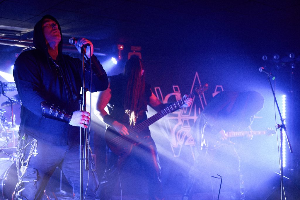 Vae Solis podczas koncertu we Wrocławiu w ramach wydarzenia „Brudna Odra” – wokalista Inferatu na pierwszym planie, w tle basista i gitarzyści zespołu oświetleni niebieskimi światłami.