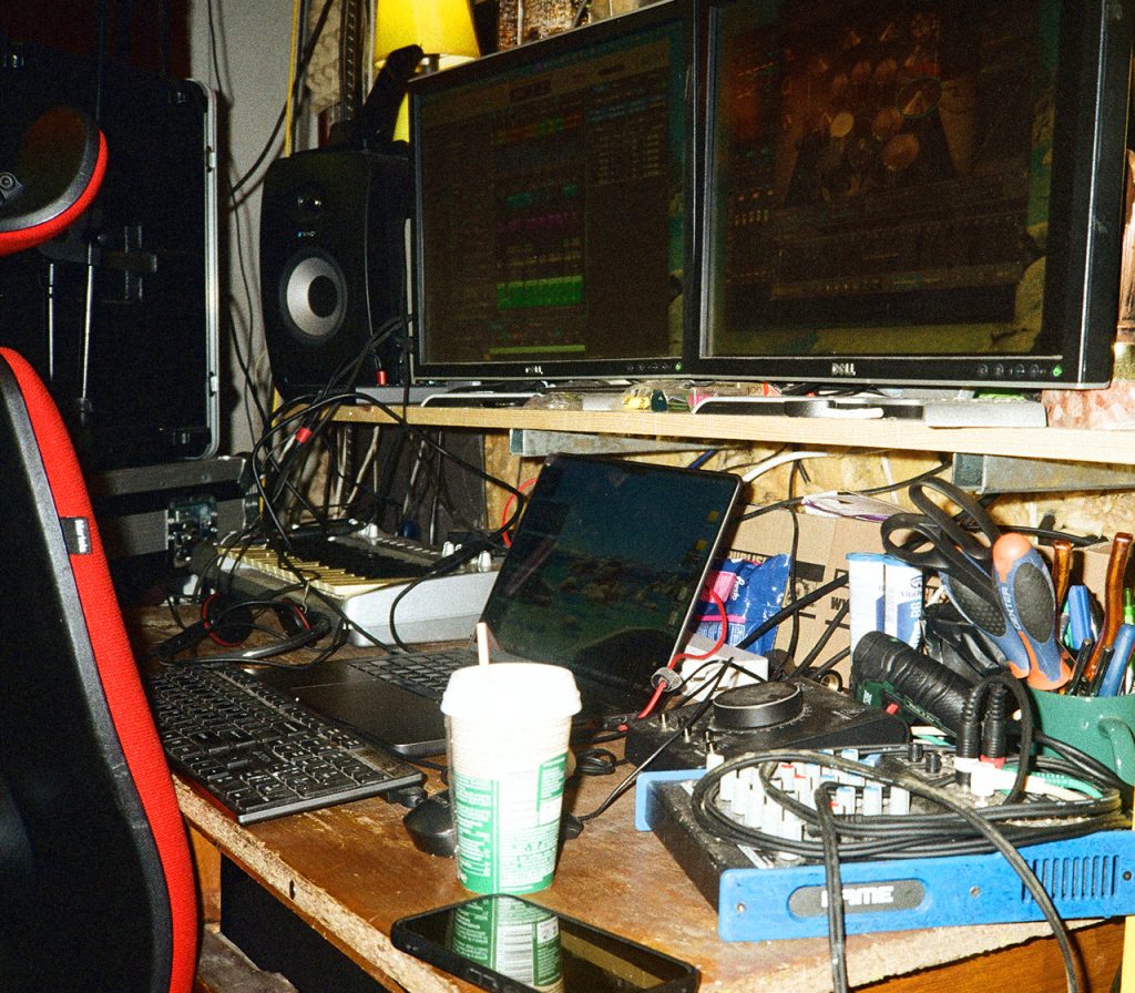 Stanowisko do pracy nad muzyką w sali prób Furydate: dwa monitory komputerowe z otwartym programem do produkcji audio, laptop, klawiatura MIDI, głośniki studyjne, tablet oraz rozbudowana plątanina kabli i sprzętu muzycznego na biurku.