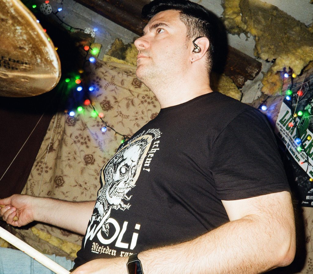 Tony, perkusista Furydate, podczas gry na perkusji w podziemnej sali prób. Na zdjęciu widać jego twarz z perspektywy bliska, słuchawkę douszną oraz pałeczkę perkusyjną w dłoni. W tle znajdują się surowe, częściowo odsłonięte ściany piwnicy, przewody i kolorowe lampki tworzące charakterystyczny klimat sali prób.