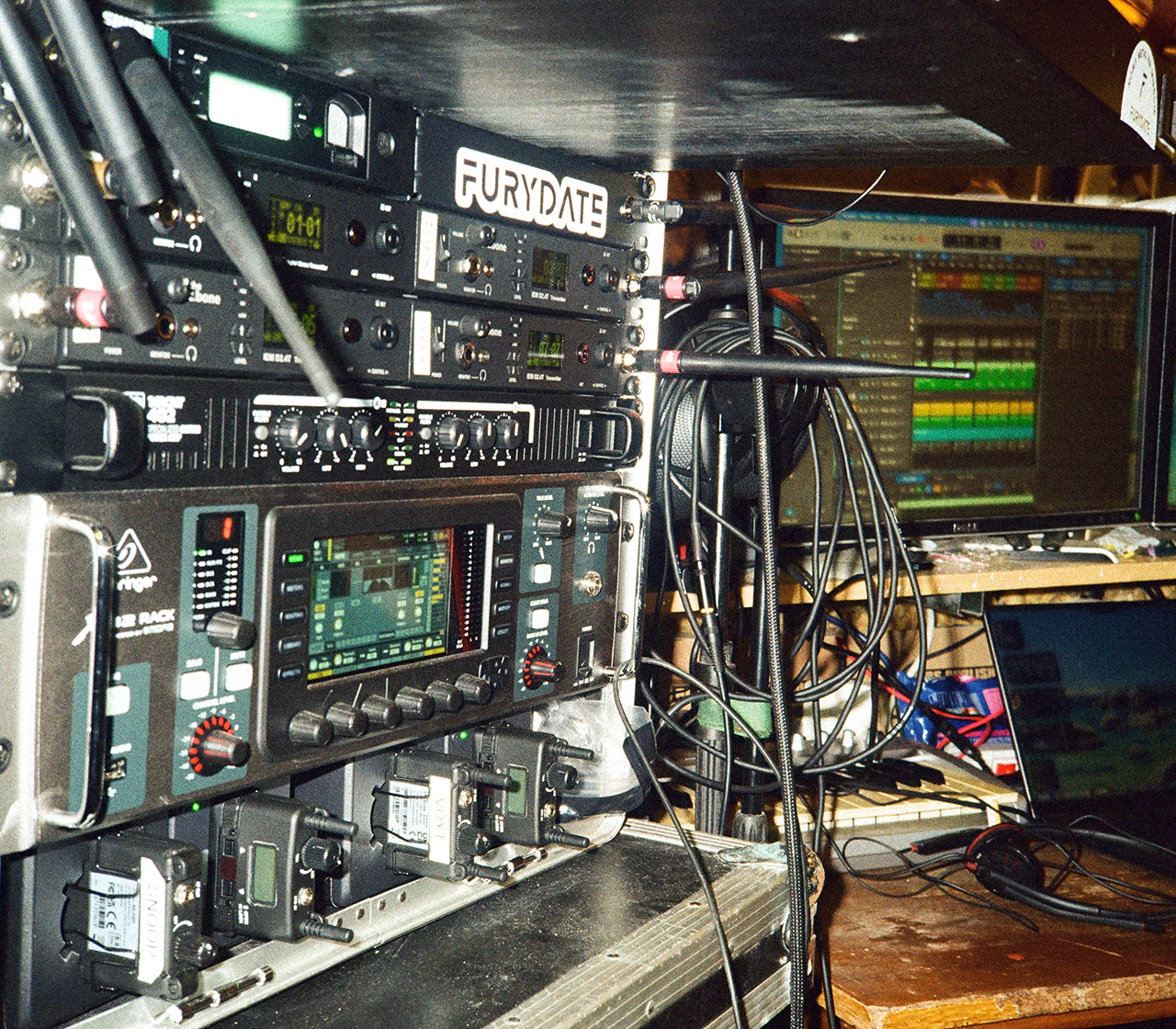 Rozbudowany rack sprzętu muzycznego Furydate w sali prób, z procesorami i urządzeniami audio, plątaniną przewodów oraz komputerowym stanowiskiem z ekranem wyświetlającym projekt muzyczny. Na jednym z urządzeń widoczne logo Furydate.