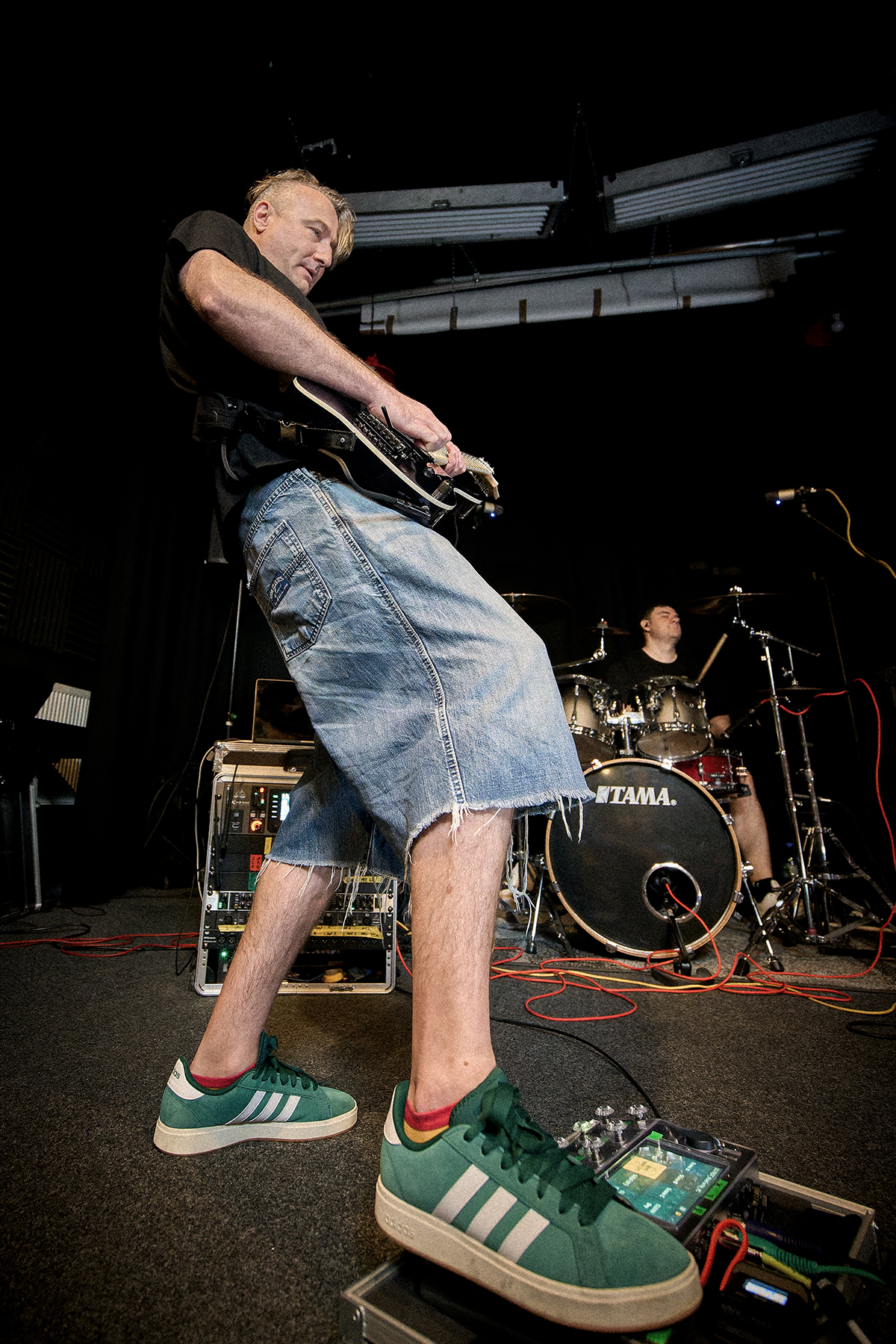 Max, gitarzysta zespołu Furydate, podczas reportażu Wajbzz gra na gitarze elektrycznej. Muzyk stoi w charakterystycznej szerokiej pozycji, z nogą opartą na pedalboardzie, a w tle znajduje się Tony za zestawem perkusyjnym. Wokół widoczne są wzmacniacze, rack ze sprzętem, mikrofony i przewody, tworzące surowy klimat sali prób.