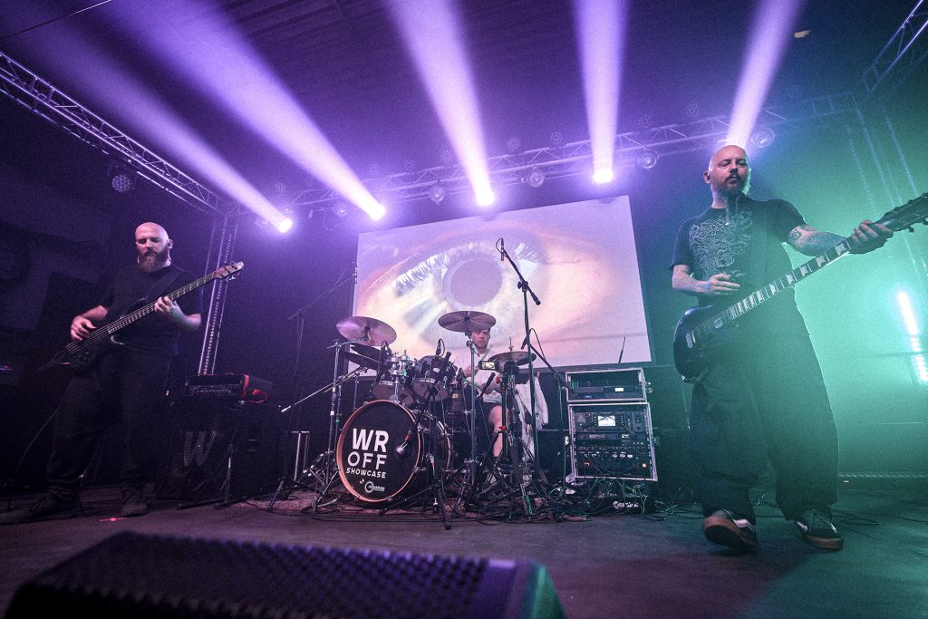 NOOMN podczas koncertu we wrocławskim klubie Łącznik – Jacek na basie, Paweł na perkusji i Kai na gitarze, na tle wizualizacji przedstawiającej ludzkie oko.